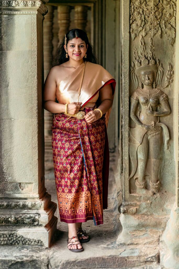 Woman wearing traditional Cambodian dress at Angkor Wat beside a carved devata figure, showing the connection between modern Khmer clothing and Angkor-period imagery.